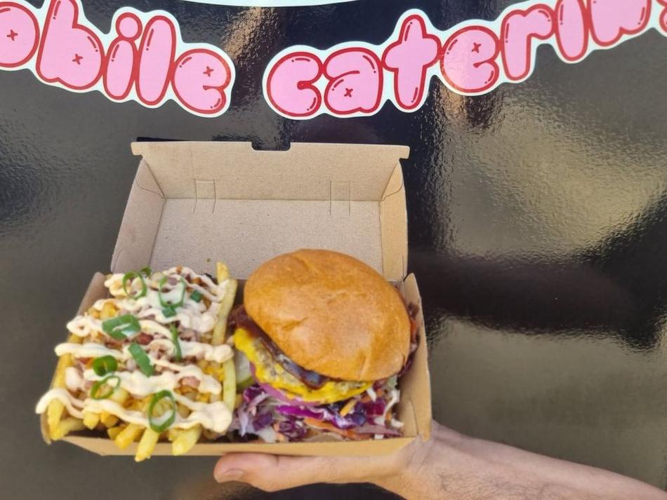 A Porkie's box holding loaded fries with bacon and shallots alongside a burger, held up against the truck